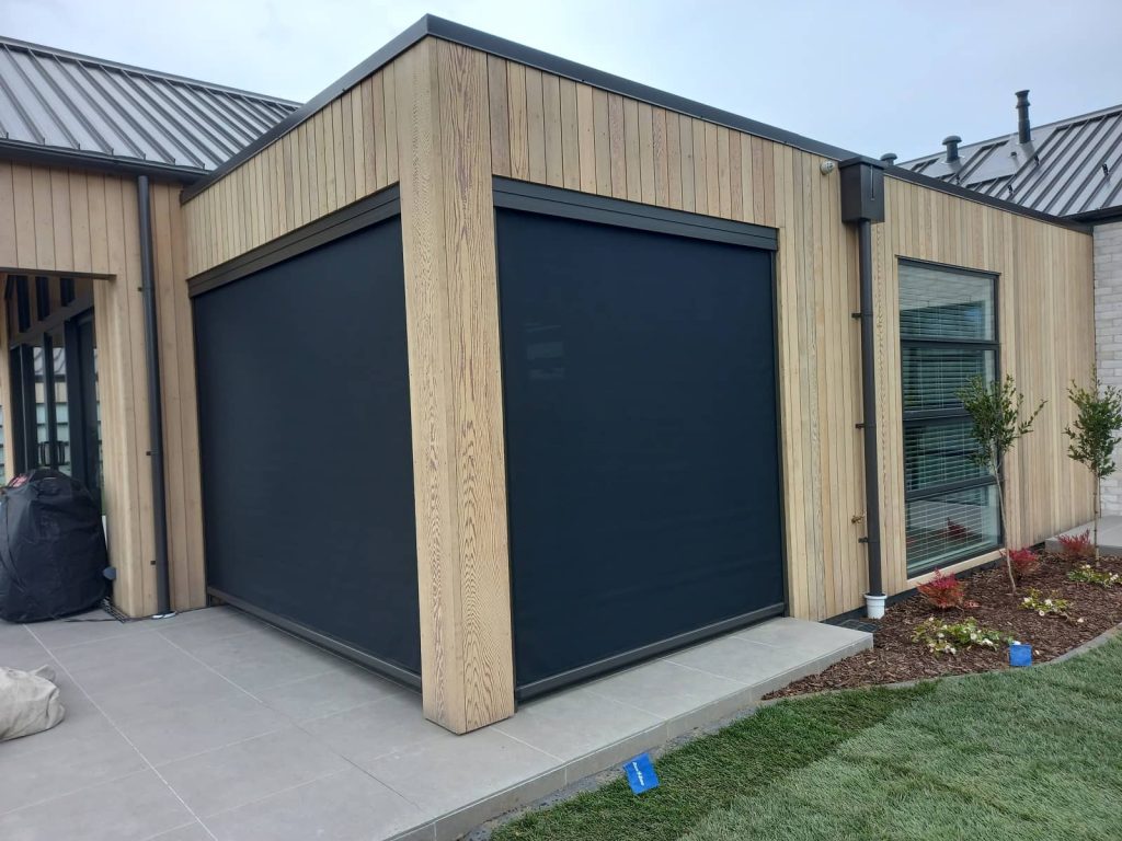 A pair of black Ziptrak blinds inset into a louvre roof with wooden cladding