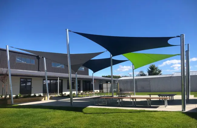 Porangahau School courtyard with modern black, grey, and green outdoor shade sails using Rainbow Shade NZ - Extreme32 covering outdoor, seating and picnic tables, under a clear blue sky