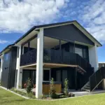 Modern two-storey beach front home with outdoor living blinds installed on an upper deck, featuring mesh outdoor blinds using Visiontex® Plus 94 Carbon Black and Ziptrak® blinds by Covered and Rainbow Shade NZ, showcasing premium outdoor screens for weather protection
