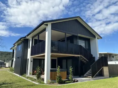 Modern two-storey beach front home with outdoor living blinds installed on an upper deck, featuring mesh outdoor blinds using Visiontex® Plus 94 Carbon Black and Ziptrak® blinds by Covered and Rainbow Shade NZ, showcasing premium outdoor screens for weather protection