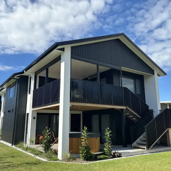 Modern two-storey beach front home with outdoor living blinds installed on an upper deck, featuring mesh outdoor blinds using Visiontex® Plus 94 Carbon Black and Ziptrak® blinds by Covered and Rainbow Shade NZ, showcasing premium outdoor screens for weather protection